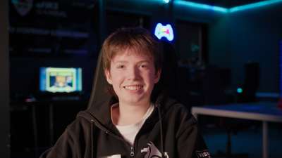 Ung gutt sitter i en gamingstol i et rom med blå belysning og datamaskiner i bakgrunnen. Han smiler og har på seg en hettegenser med logoer.