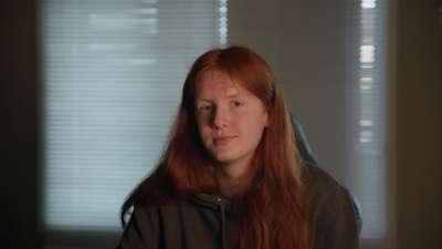 En ung guttmed langt rødt hår sitter i en gamingstol foran et vindu med persienner. Personen har på seg en mørk hettegenser og ser rolig mot kamera.
