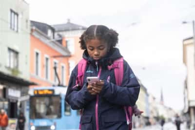 Jente i tiårsalederen med skolesekk på ryggen. Hun er i et bymiljø, med trikk i bakgrunnen. Jenta ser ned på mobiltelefonen sin, som hun holder med begge hender.